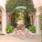 Gray Malin Via Parigi Worth Avenue, Palm Beach features two peach bicycles with baskets beneath a "VIA PARIGI" archway, lush plants, and pink flowers. Archival luster paper print in white frame, 24"x36", by Gray Malin.