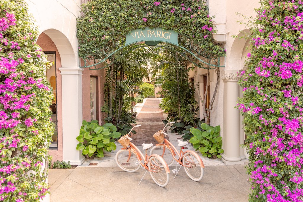 Gray Malin Via Parigi Worth Avenue, Palm Beach features two peach bicycles with baskets beneath a "VIA PARIGI" archway, lush plants, and pink flowers. Archival luster paper print in white frame, 24"x36", by Gray Malin.