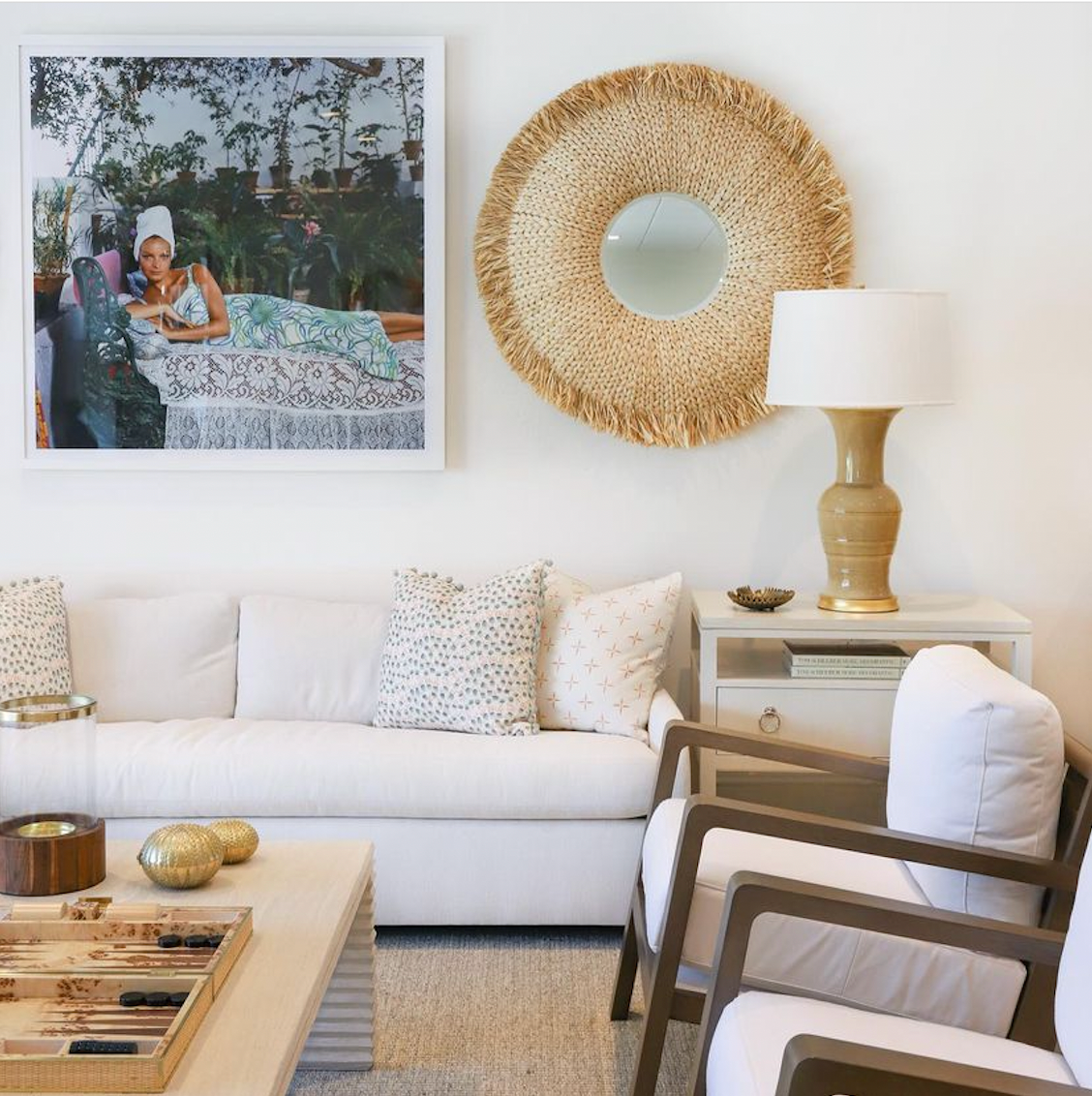 A living room furnished with a white couch, patterned pillows, wooden chairs, and an eclectic décor showcasing the Made Goods Casey Round Natural Raffia Mirror. A table lamp brightens the space, paired with wall art of a lounging woman.