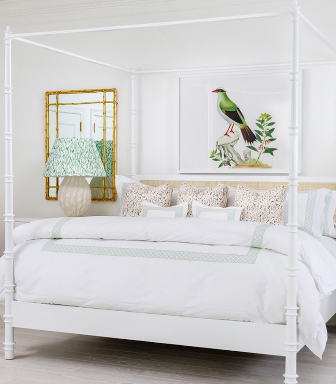 A white four-poster bed features Sorella Glenn’s Feather Ikat Shade in Spearmint bedding in a bright bedroom, paired with a green lamp. Above the bed are a gold-framed mirror and bird artwork.