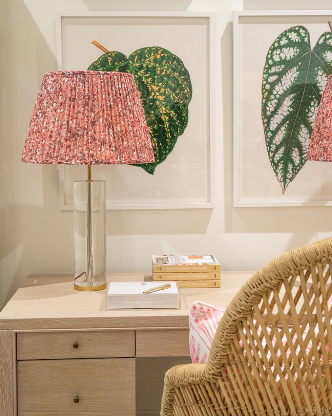 A desk with a clear lamp featuring a pink patterned shade, a notepad, books, and a pen, highlighted by Ornis Gallery's English Leaves Print 8213 on the wall in Larson-Juhl frames, with a wicker chair in the foreground.