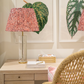 A wicker chair faces a light wooden desk with a pink patterned lamp, books, papers, and a notebook; two Ornis Gallery “English Leaves Print 8259” botanicals adorn the wall in Larson-Juhl frames.