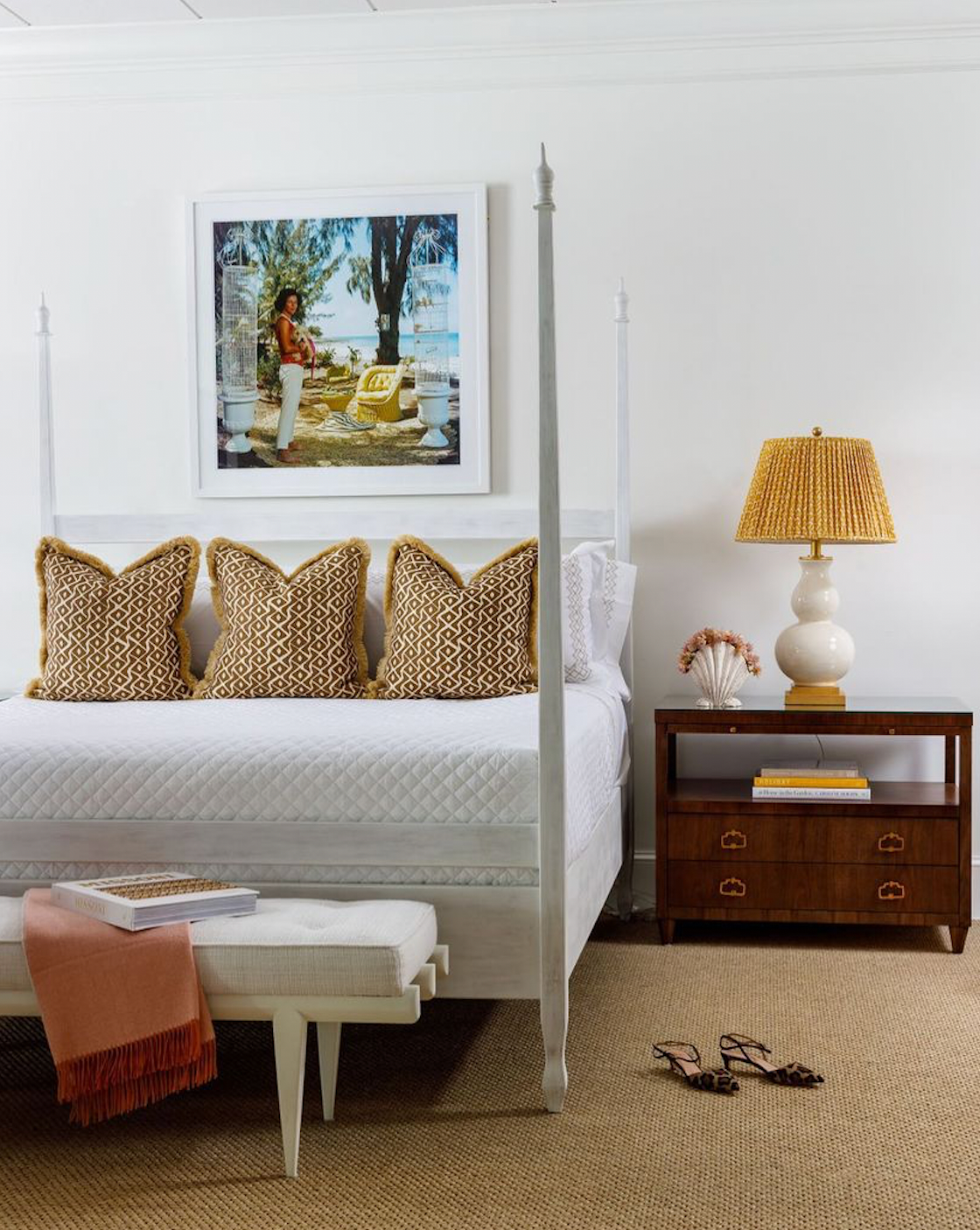 A bedroom featuring Noir's Four Poster Whitewash Venice Bed, Queen, with patterned pillows, a mahogany nightstand and lamp, a bench at the bed's foot, slippers on the floor, and framed artwork on the wall.