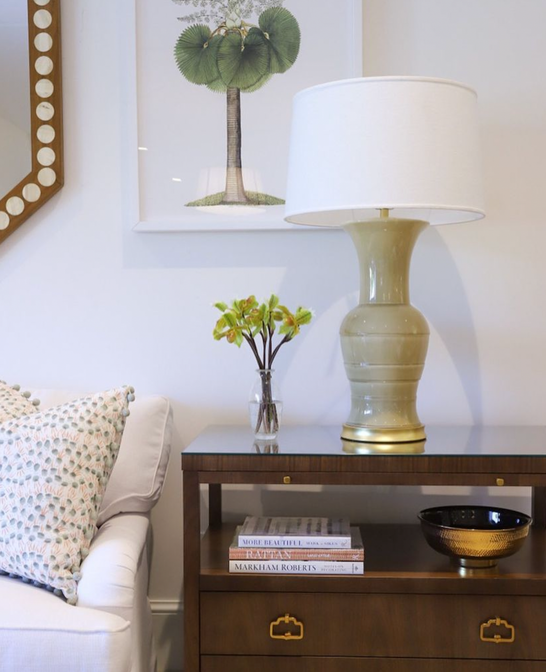 A side table with Hwang Bishop's Koi Lamp in Cream Ceramic with a warm silver base, a floral vase, books, and a decorative bowl sits by a white sofa; framed art and a mirror hang on the wall behind.