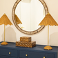 A Hyson Round Mirror by Currey and Company adorns the wall above a blue dresser with gold knobs. Flanked by two wicker lamps with conical shades, patterned decorative boxes complete the elegant, stylish setup.