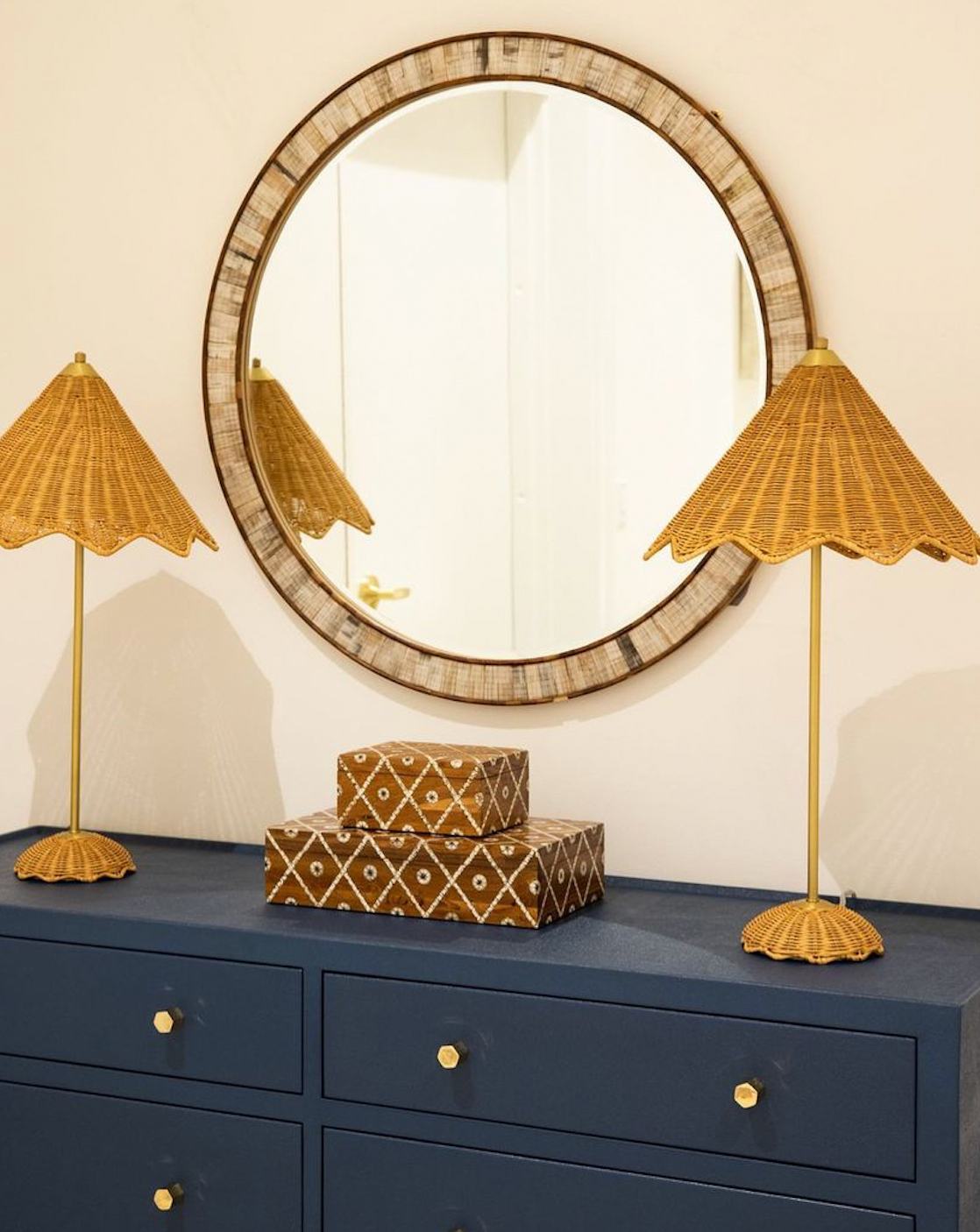 A Hyson Round Mirror by Currey and Company adorns the wall above a blue dresser with gold knobs. Flanked by two wicker lamps with conical shades, patterned decorative boxes complete the elegant, stylish setup.