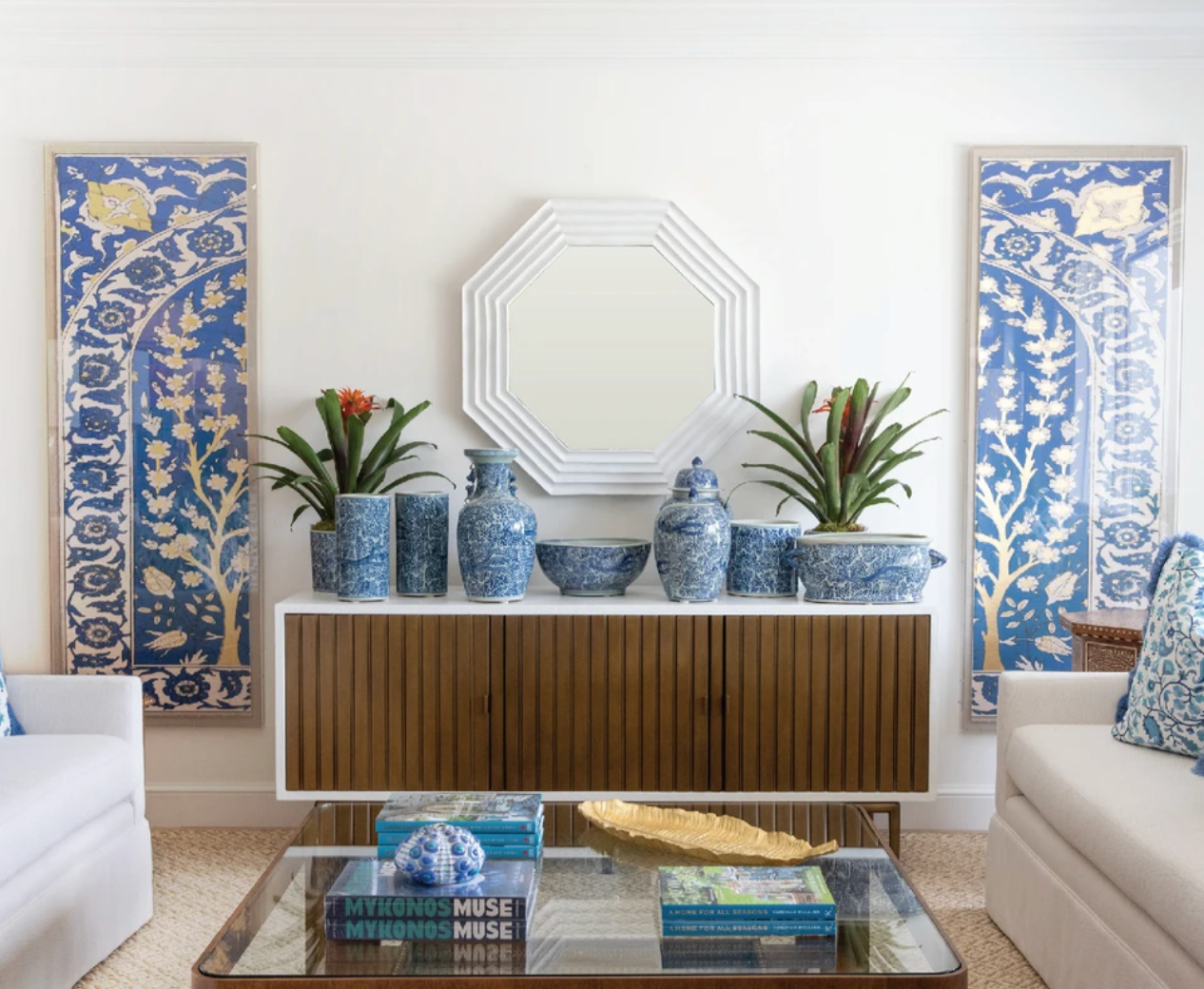 In the living room, the Naples Cabinet by Mr Brown, adorned with aged brass doors, is topped with blue and white vases beneath a central mirror. Two framed blue artworks and potted plants accent the space. Facing one another are two white sofas finished in smooth gesso, creating an inviting ambiance.