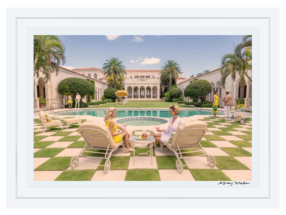 Two people relax on lounge chairs by a pool in an opulent courtyard with palm trees and a villa, evoking the retro luxury of Gray Malin’s "Poolside at La Follia, Palm Beach" series.