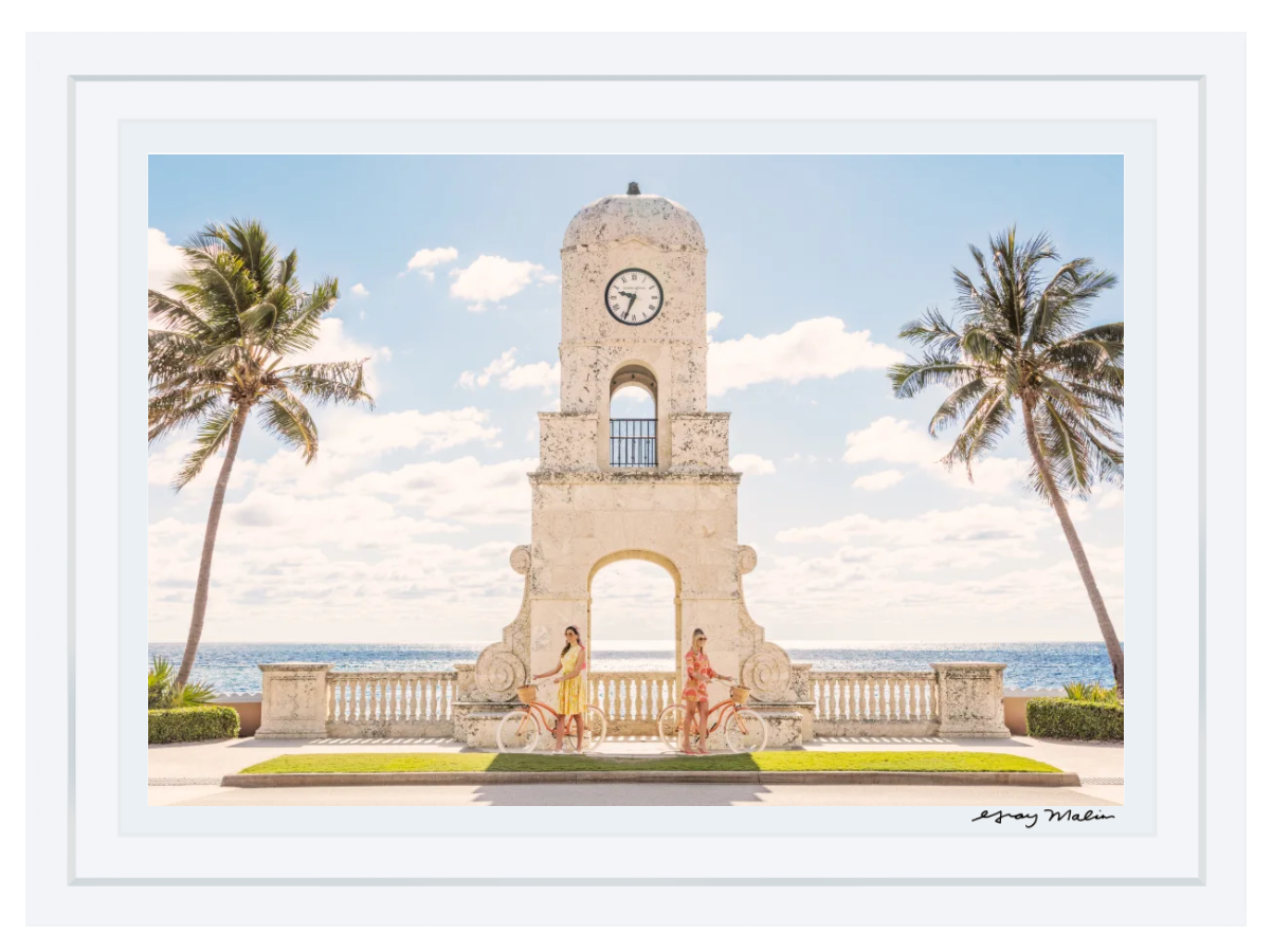 Inspired by Gray Malin’s Worth Avenue Clock Tower (29.5" x 41.5", White Frame), two cyclists pass the iconic Palm Beach landmark, palm trees and the ocean beneath a brilliant sky—a scene reflecting Gray Malin’s signature style.