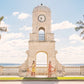 Two people cycle past Palm Beach's Worth Avenue Clock Tower, palms swaying under a bright sky, in a scene recalling Gray Malin’s iconic style—mirrored in the Gray Malin Worth Avenue Clock Tower print, White Frame, 29.5" x 41.5".