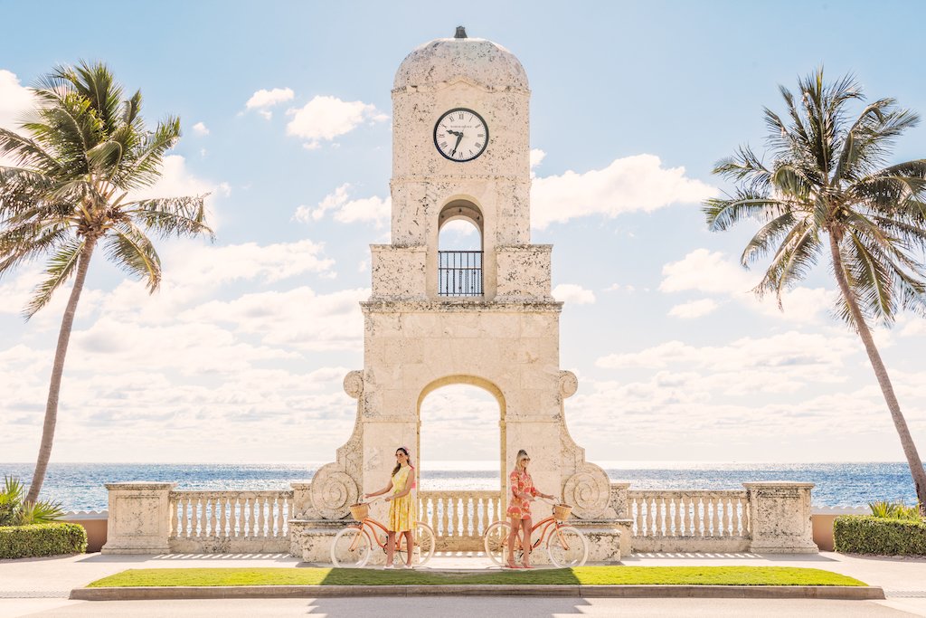Two people cycle past Palm Beach's Worth Avenue Clock Tower, palms swaying under a bright sky, in a scene recalling Gray Malin’s iconic style—mirrored in the Gray Malin Worth Avenue Clock Tower print, White Frame, 29.5" x 41.5".